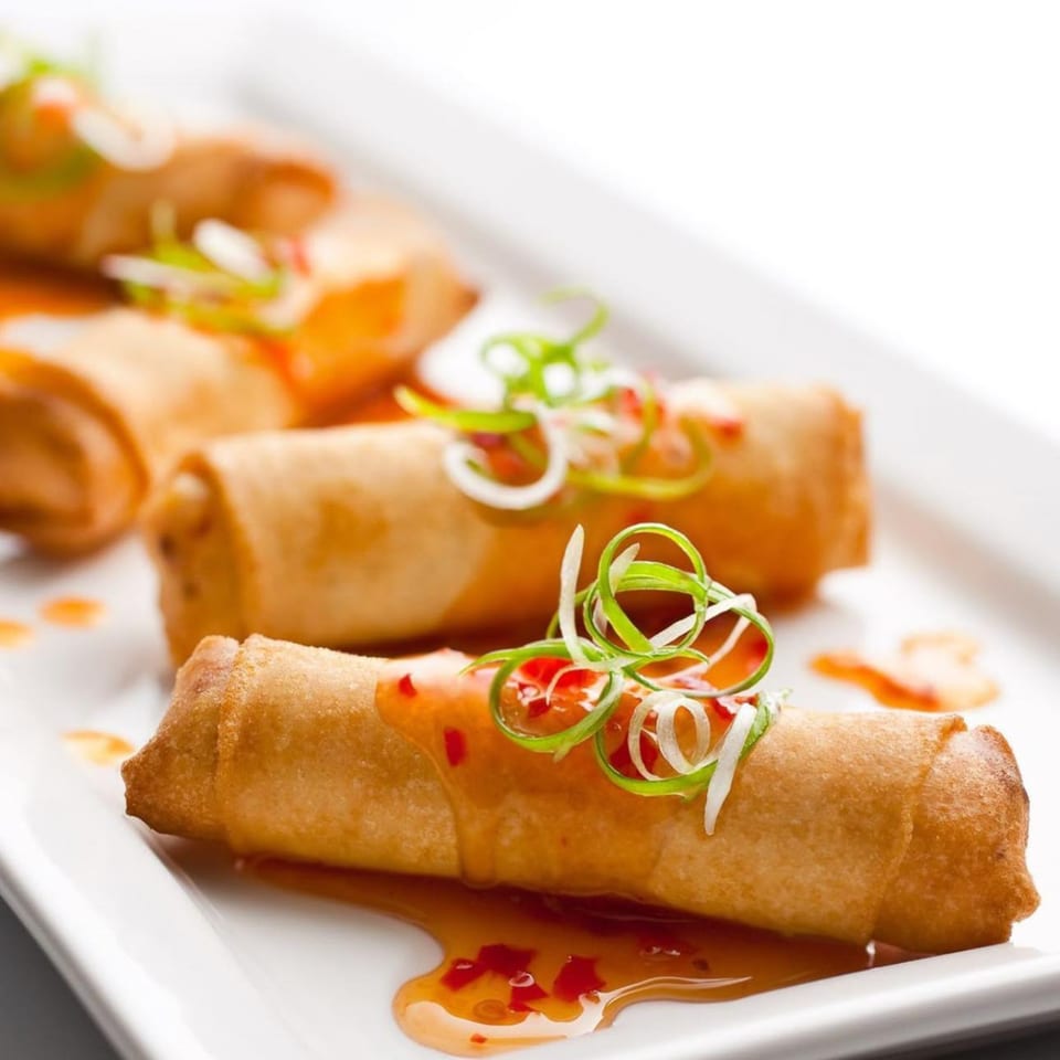 Crispy Vietnamese spring rolls drizzled with sweet chili sauce and garnished with green onions on a white plate.