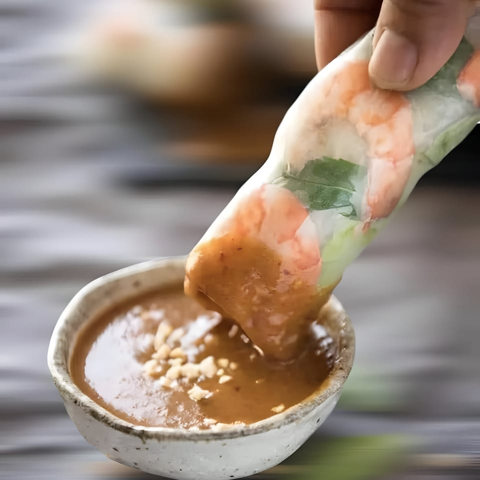 A hand dips a fresh Vietnamese spring roll with shrimp and greens into a peanut sauce.