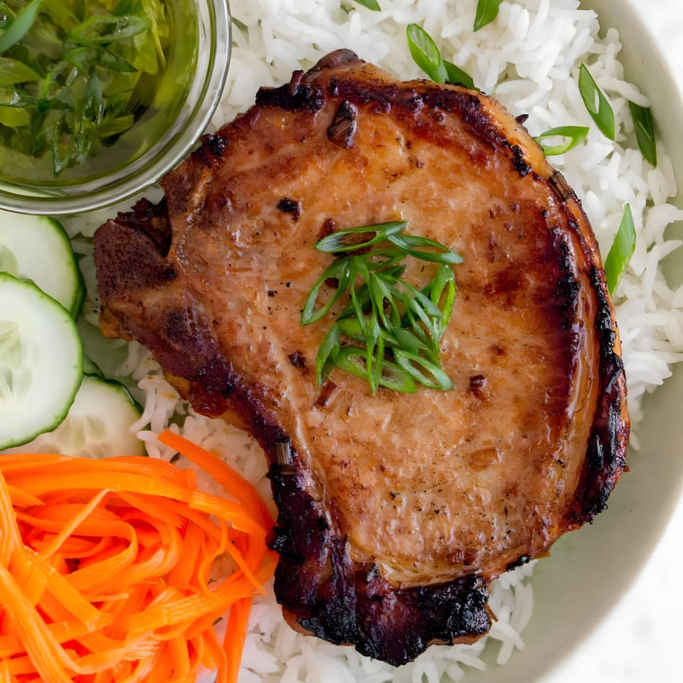 Grilled pork chop with rice, shredded carrots, cucumber slices, and a side of green sauce.